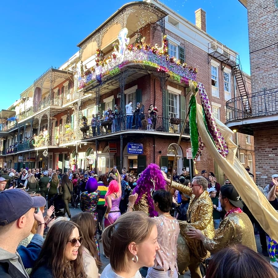 A vibrant Mardi Gras parade fills a street in New Orleans, with colorful costumes and festive decorations on the balconies above.