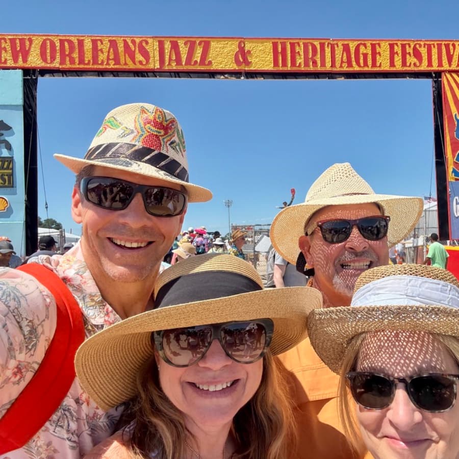 A group of four friends poses in front of the New Orleans Jazz & Heritage Festival sign, wearing sun hats and sunglasses.