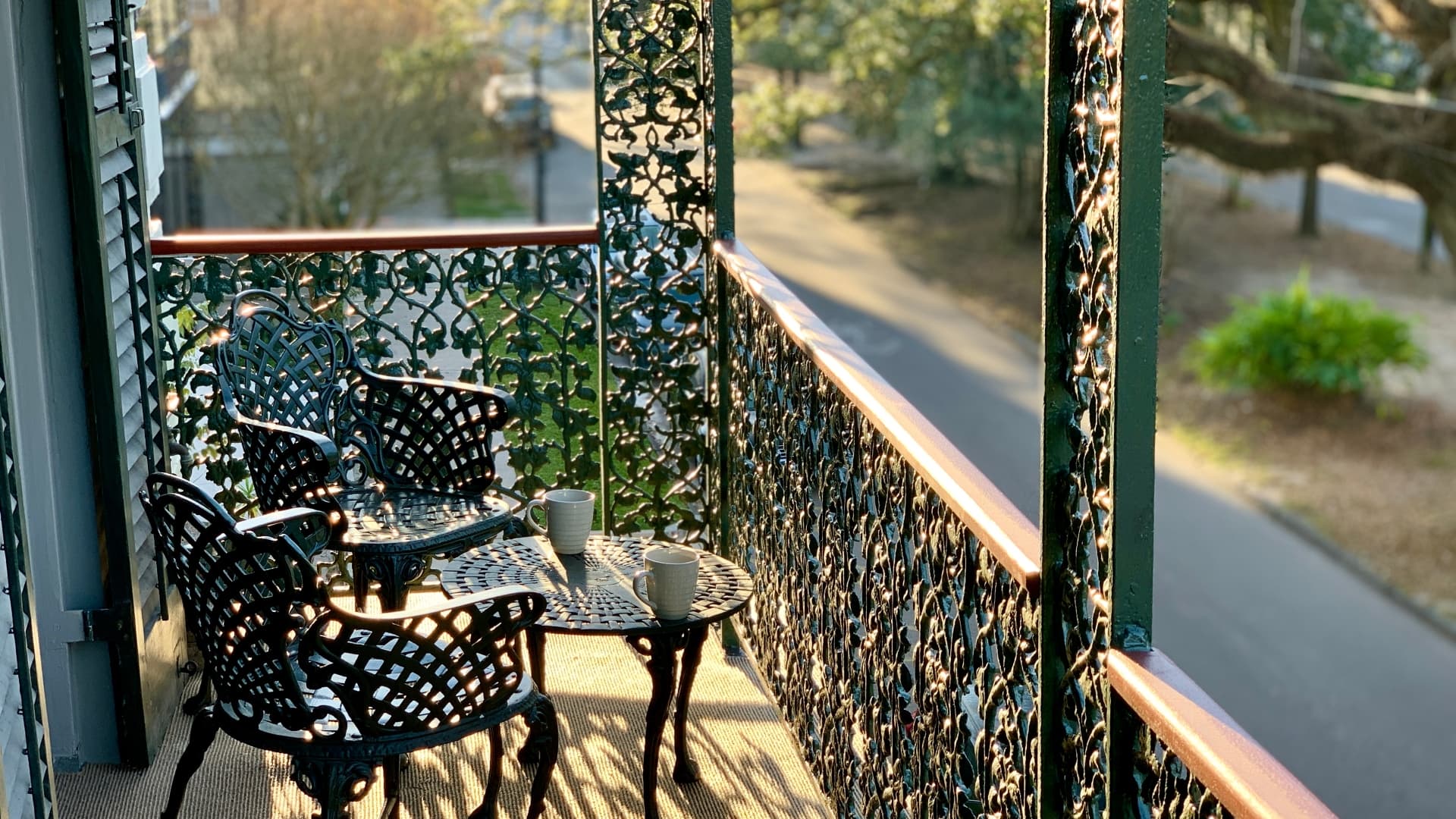A sunlit balcony with ornate black chairs and a small table, featuring two cups.